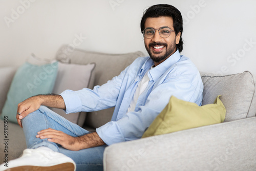 Happy confident handsome millennial arab guy in glasses with beard sits on sofa, looking at camera