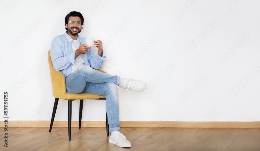 Cheerful millennial arab male in casual, glasses with beard sits on armchair with cup of hot drink