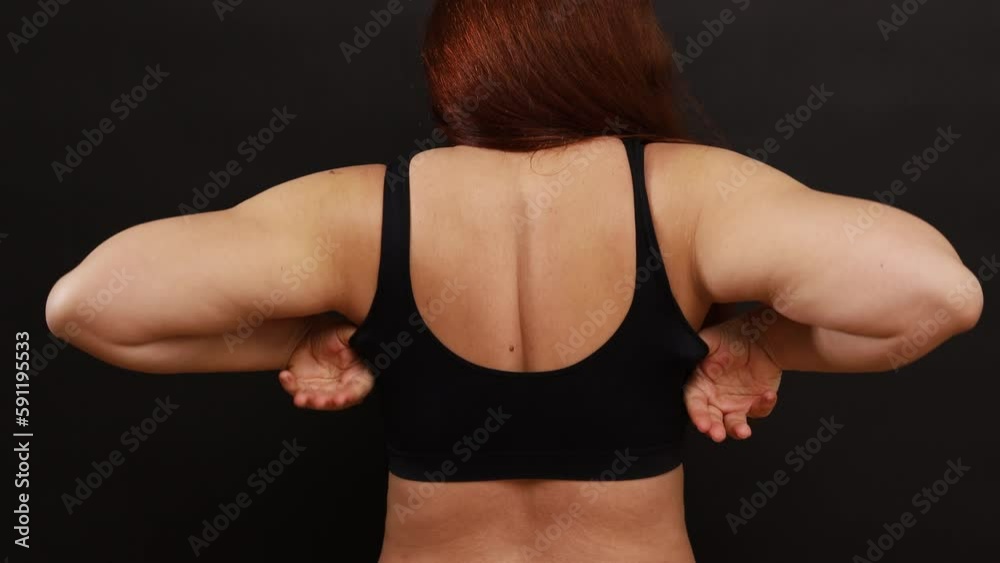 Back view of plump woman with long red hair adjusting straps, trying on black sports bra, standing on black background. Overweight, healthcare, body positive, weight loss, underwear, liposuction.