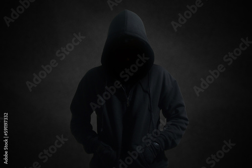 Mysterious man wearing black hoodie standing against dark background. Hacker, crime, and cyber security concept.
