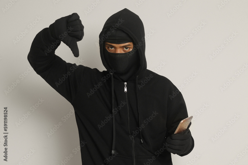 Portrait of mysterious man wearing black hoodie and mask doing hacking activity on mobile phone, hacker holding a smartphone. Isolated image on gray background Portrait of mysterious man wearing black hoodie and mask doing hacking activity on mobile phone, hacker holding a smartphone. Isolated image on gray background
