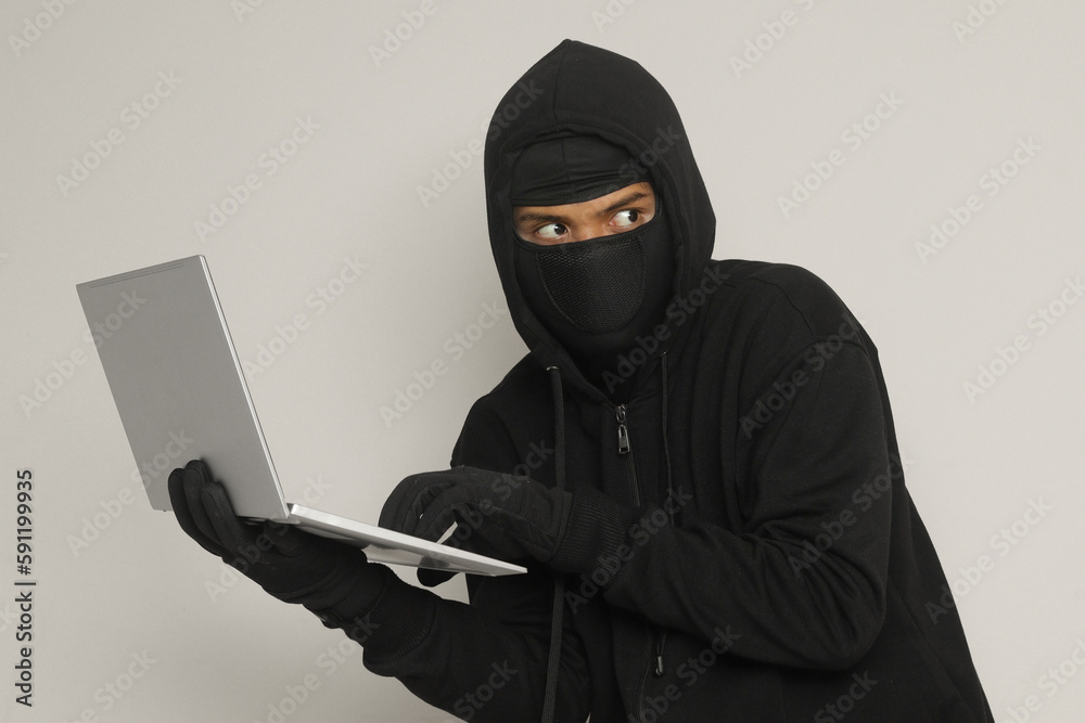 Portrait of mysterious man wearing black hoodie and mask doing hacking activity on laptop, hacker holding a personal computer. Cyber security concept. Isolated image on gray background Portrait of mysterious man wearing black hoodie and mask doing hacking activity on laptop, hacker holding a personal computer. Cyber security concept. Isolated image on gray background