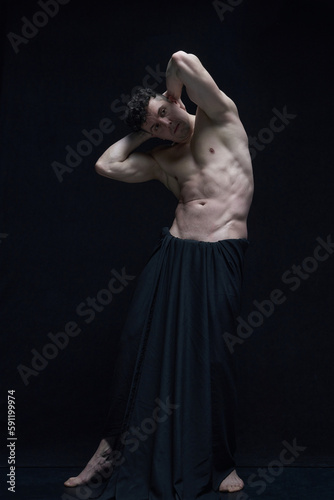 Full-length portrait of mature, muscular, handsome, shirtless man posing in dark fabric against black studio background. Concept of male body aesthetics, men's beauty, inspiration, art