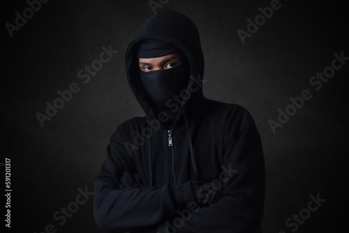 Mysterious man wearing black hoodie and mask standing against dark background, looking at camera. Dramatic low light portrait
