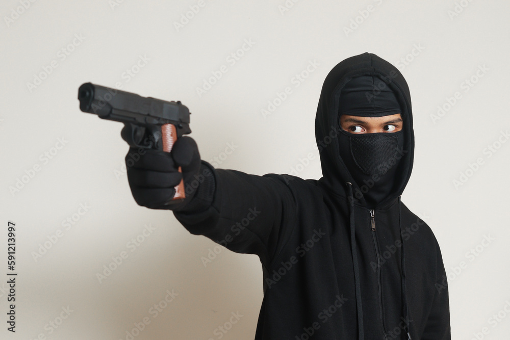 Mysterious man wearing black hoodie and mask holding a pistol, shooting with a gun. Isolated image on gray background Mysterious man wearing black hoodie and mask holding a pistol, shooting with a gun. Isolated image on gray background