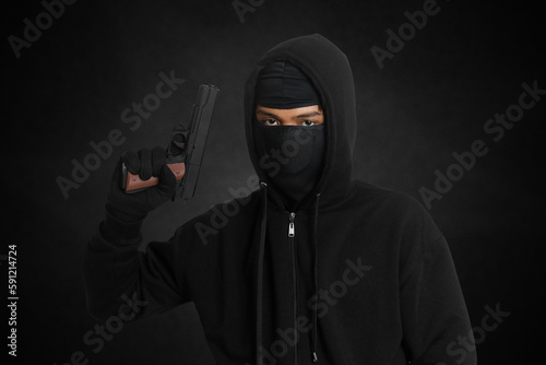 Fototapeta Naklejka Na Ścianę i Meble -  Mysterious man wearing black hoodie and mask holding a pistol, shooting with a gun. Isolated image on dark ambient background