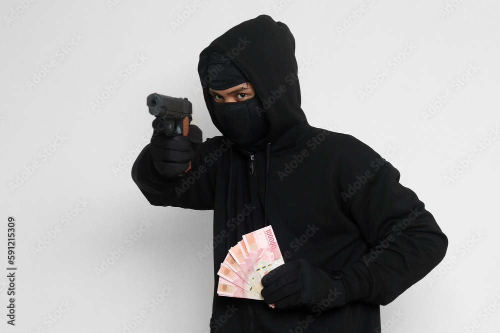 Portrait of mysterious man wearing black hoodie and mask doing shooting with gun while holding one hundred thousand rupiah banknote, extorting money. Isolated image on gray background Portrait of mysterious man wearing black hoodie and mask doing shooting with gun while holding one hundred thousand rupiah banknote, extorting money. Isolated image on gray background