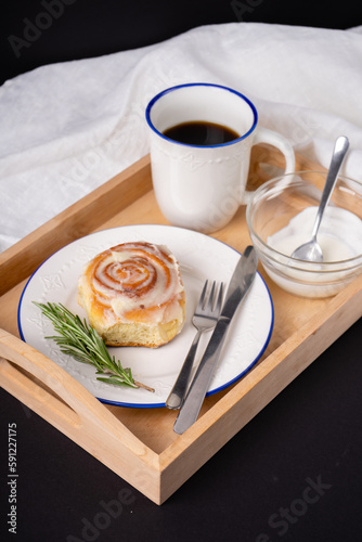 cinnamon roll with rosemary on a white plate, a cup of coffee and cream on a wooden tray