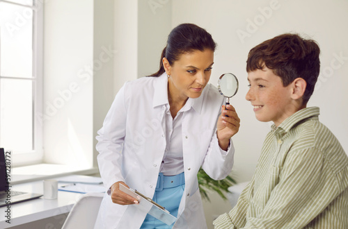 Doctor examines skin on child s face. Professional dermatologist uses loupe to investigate growth on face of happy freckled little boy and eliminate risk of cancer disease diagnosis. Checkup concept