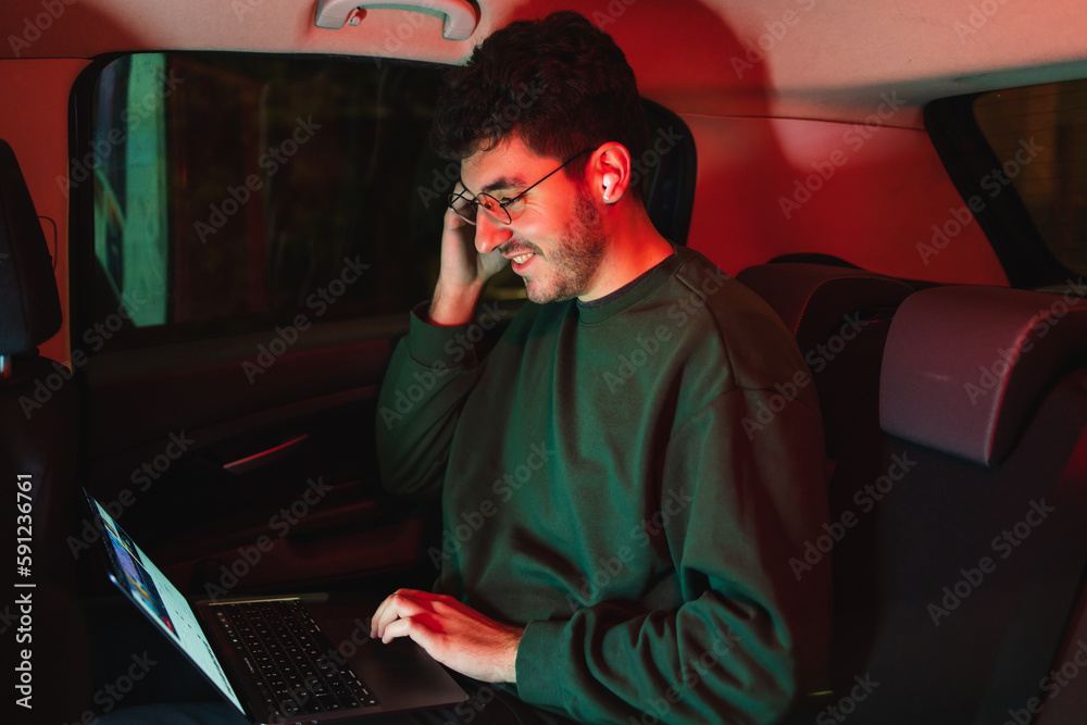 One young man is working from car in backseat while using laptop and mobile phone to work during the night One young man is working from car in backseat while using laptop and mobile phone to work during the night