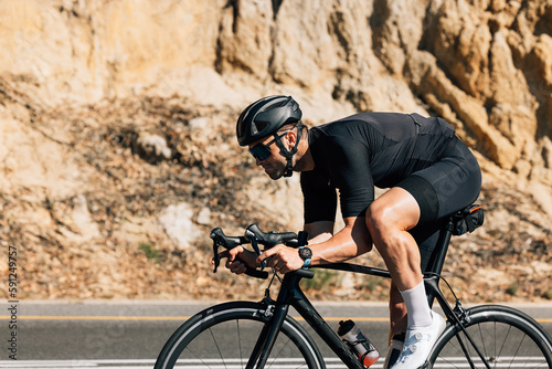 Professional road bike rider in black sportswear. Side view of a young male riding bicycle against a mountain.