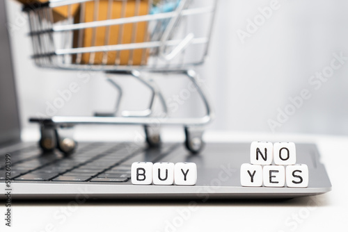 Buy yes or no words on cubes laying on the laptop and a toy shopping cart with carton boxes serves as the background.