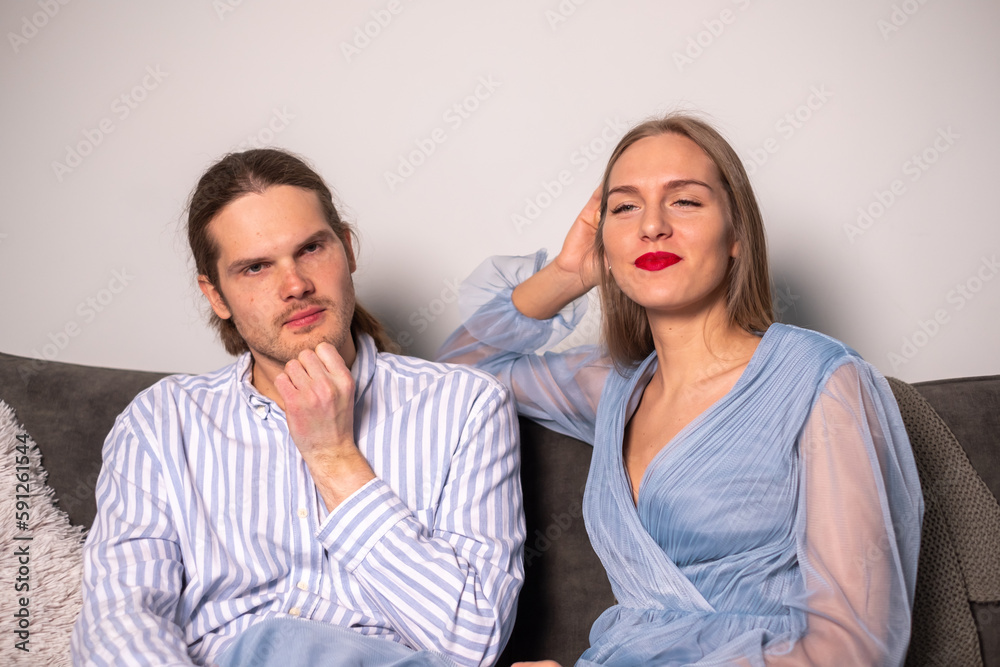 A thoughtful man in a striped shirt and a beautiful smiling woman with long hair, red lips, a stylish light blue dress are sitting on a sofa and looking at the camera. A thoughtful man in a striped shirt and a beautiful smiling woman with long hair, red lips, a stylish light blue dress are sitting on a sofa and looking at the camera.