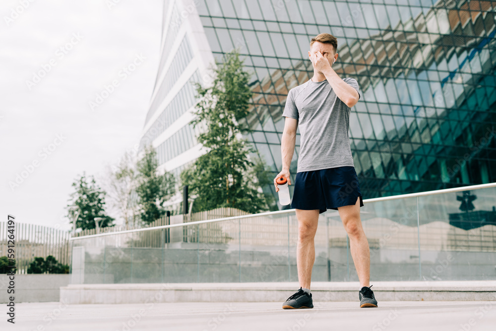 Tired Young man runner drinking water, relaxing after sport training. Holding water bottle while doing fitness workout in summer city urban street, cloudy sky Tired Young man runner drinking water, relaxing after sport training. Holding water bottle while doing fitness workout in summer city urban street, cloudy sky