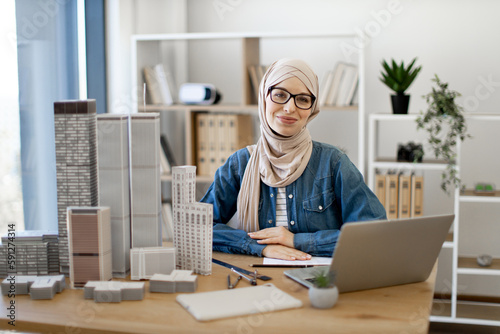 Young muslim woman wearing hijab and spectacles sitting at office desk in real estate agency. Efficient realtor using wireless technologies while arranging purchase of residential properties indoors.
