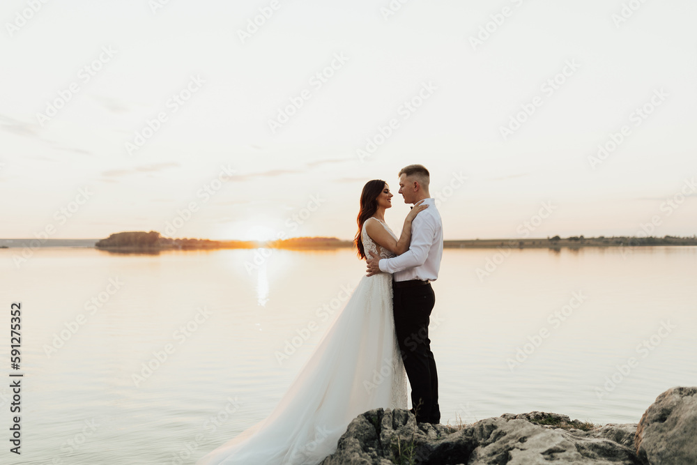 Fototapeta premium Groom and bride standing on the rocks. They hugging and looking each other, just married couple. Outdoor wedding ceremony on the seashore.