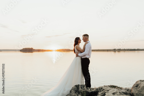 Groom and bride standing on the rocks. They hugging and looking each other, just married couple. Outdoor wedding ceremony on the seashore.