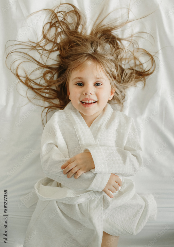 Joyful girl in a white bathrobe lies in bed, looks at the camera. - obrazy, fototapety, plakaty 