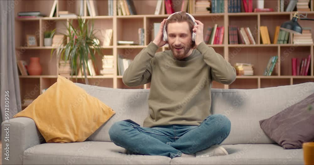 Attractive Caucasian man wearing headphones and resting in library. Comfortably sitting in lotus pose on couch and meditating, listening relaxing sounds and calming music. Yoga, wellness concept. Attractive Caucasian man wearing headphones and resting in library. Comfortably sitting in lotus pose on couch and meditating, listening relaxing sounds and calming music. Yoga, wellness concept.