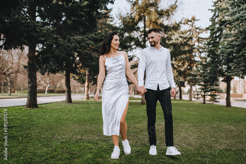 A bearded young groom and a beautiful smiling bride in a white brunette dress are walking in the park outdoors, holding hands. Wedding photography of happy, cheerful newlyweds, portrait.