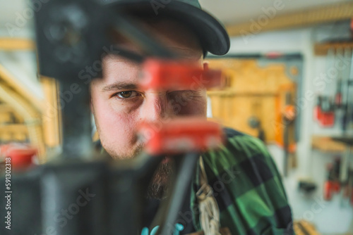 Adult male carpenter looking through red and black bar clamp and blinking at camera. Blurred wood workshop interior. High quality photo © PoppyPix