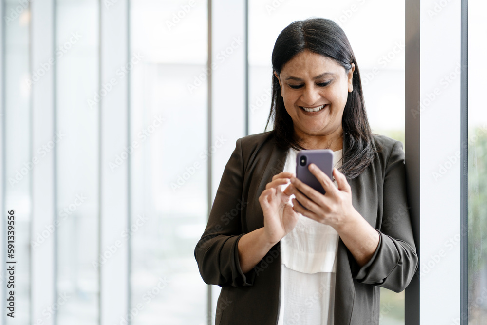 Smiling mature Asian woman holding mobile phone and checking social media content, scrolling news feed standing in modern office. Indian businesswoman using smartphone shopping online. Technology Smiling mature Asian woman holding mobile phone and checking social media content, scrolling news feed standing in modern office. Indian businesswoman using smartphone shopping online. Technology