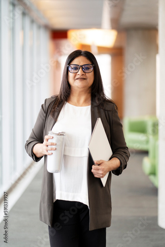 Confident Indian woman, 50 years old entrepreneur, business owner, executive director, recruiter, influencer, smiling looking at camera standing with laptop in a financial building photo