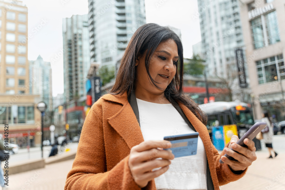 Fototapeta premium Mature Asian woman using smartphone and credit card, transferring money, standing on the city street