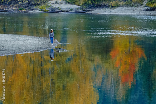 Woman standing on the bank of calm river or lake with reflection of fall thees in the water.