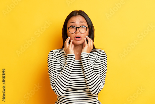 Portrait of suprised woman on yellow background, she is in black-and-white sweatshirt and holds hands up to her cheeks, copy space, high quality photo photo