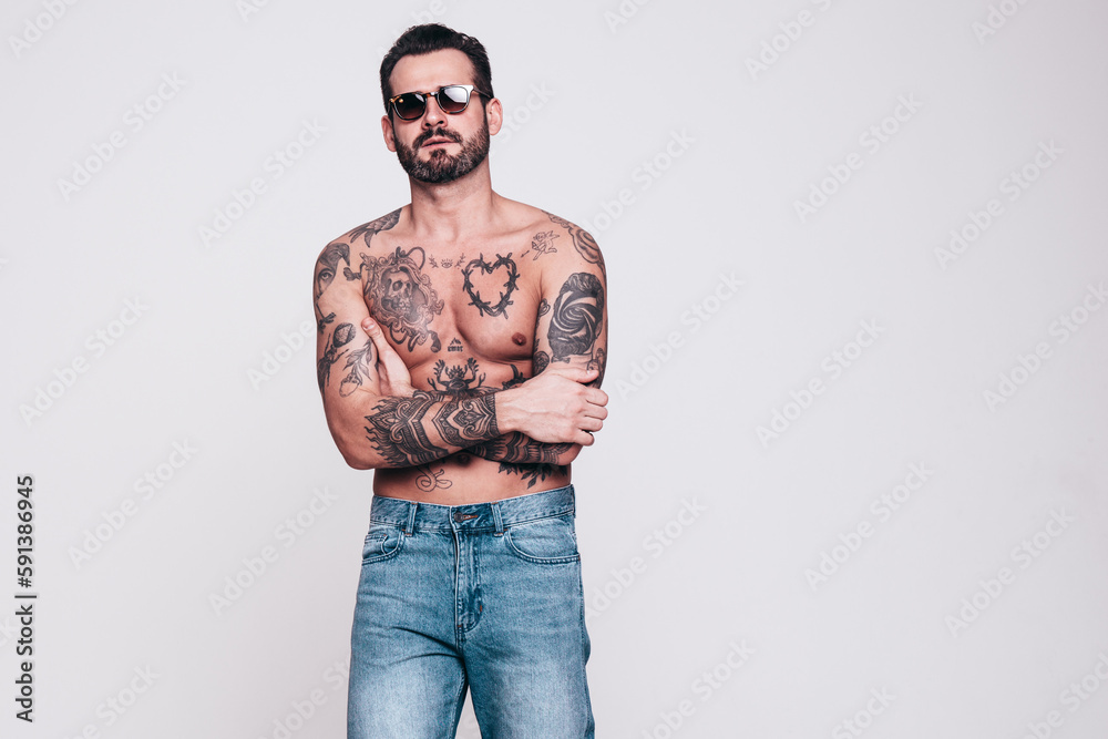 Fototapeta premium Portrait of handsome confident stylish hipster lambersexual model in jeans. Sexy modern man. Naked torso with tattoos. Fashion male posing in studio. Isolated on white. In sunglasses