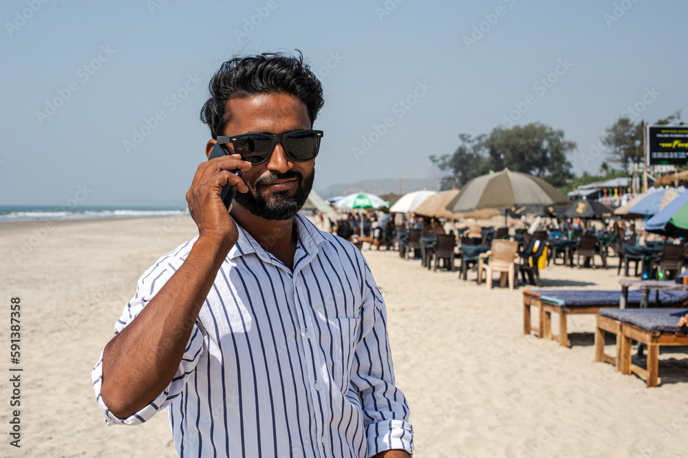 Smiling mature dark skin businessman with black hair in sunglasses, in shirt stand on sandy seashore, talk on smartphone