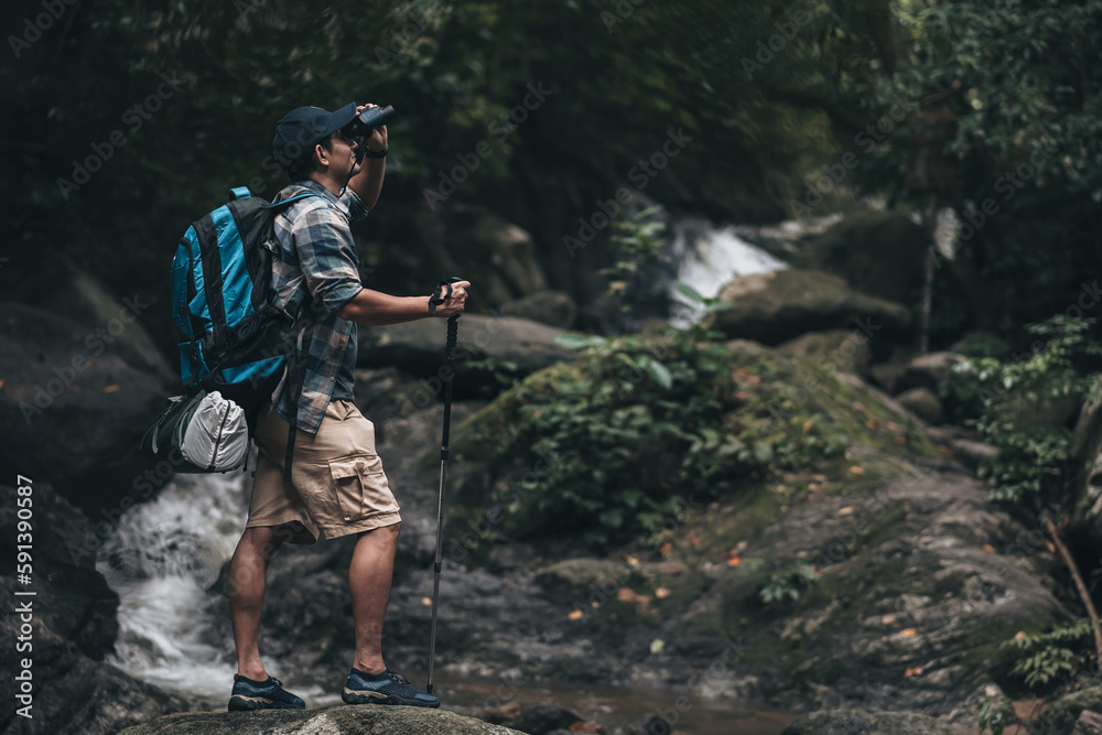 Hikers standing on the rock and use binocular to see animals and view landscape  with backpacks and background waterfall in the forest. hiking and adventure concept.