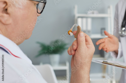 Senior patient holding a hearing aid in his hand. Deaf old man looking at a small modern medical hearing device that the doctor gave him. Close up shot. Medicine, hearing loss, treatment concept