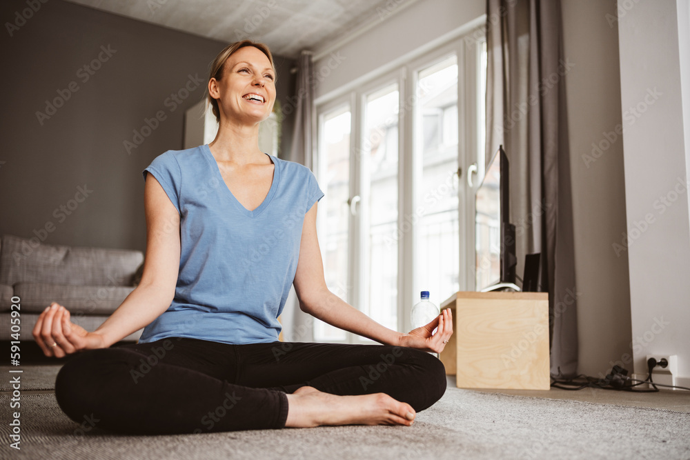 Young blonde woman sitting on the floor and practicing yoga in her apartment, laughing and looking forward