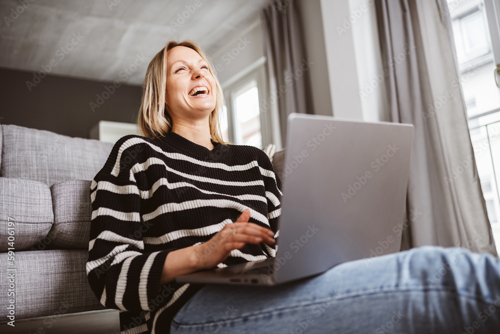 Naklejka premium Young woman sitting on the floor, laughing and looking up at the ceiling with her laptop