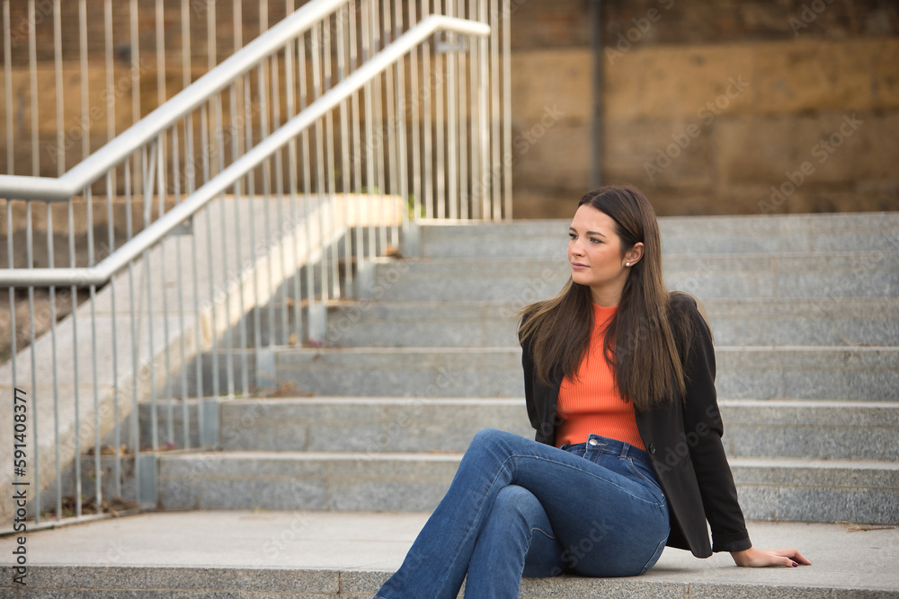 Young and beautiful woman with straight brown hair, jacket and jeans, sitting on a staircase, looking lost in infinity. Concept fashion, beauty, trend, millennial.