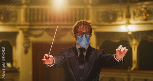Cinematic Close Up of Conductor with Medical Mask Directing Symphony Orchestra with Performers Playing on Stage During Music Concert. Professional Conductor Leading Musicians in Classic Theater