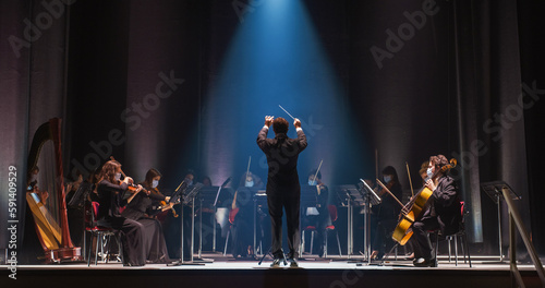 Cinematic Shot of an Orchestra on a Classic Theatre Stage:  Professional Conductor Directing Symphony Orchestra with Performers Playing Violins, Cellos, and Trumpets During Music Concert photo
