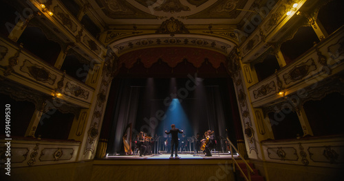 Wide shot of an Orchestra on a Classic Theatre Stage:  Professional Conductor Directing Symphony Orchestra with Performers Playing Violins, Cellos, and Trumpets During Music Concert. Audience's POV