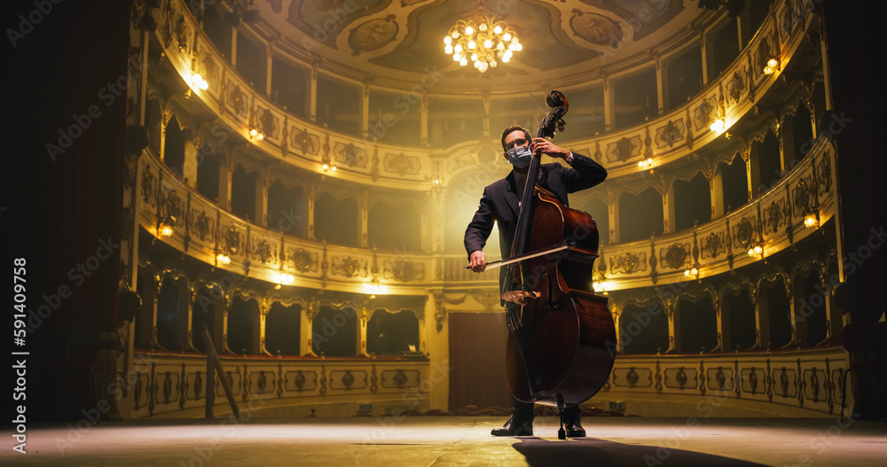 Cinematic shot of Male Cellist Wearing Protective Medical Mask, Playing Cello Solo on an Empty Classic Theatre Stage with Dramatic Lighting. Musician Rehearsing Before the Start of a Show.