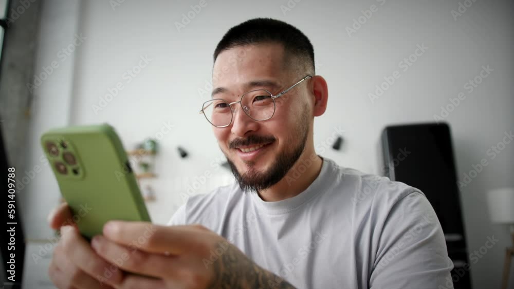 Smiling Man Texting Using Smartphone At Home. Asian man chatting online holding mobile phone in hands sitting alone. Close up. Smiling Man Texting Using Smartphone At Home. Asian man chatting online holding mobile phone in hands sitting alone. Close up.