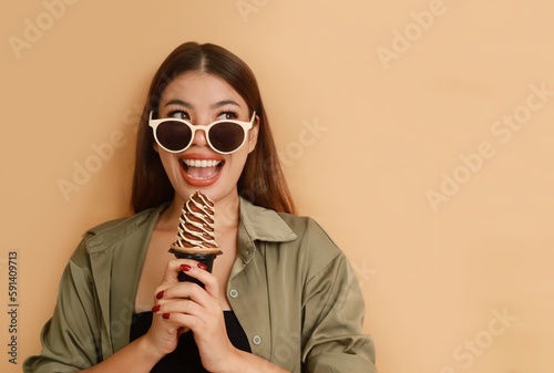 Beautiful young Asian in casual shirt holding big gelato standing over beige background isolated, copy space. Girls Summer joyful concept.