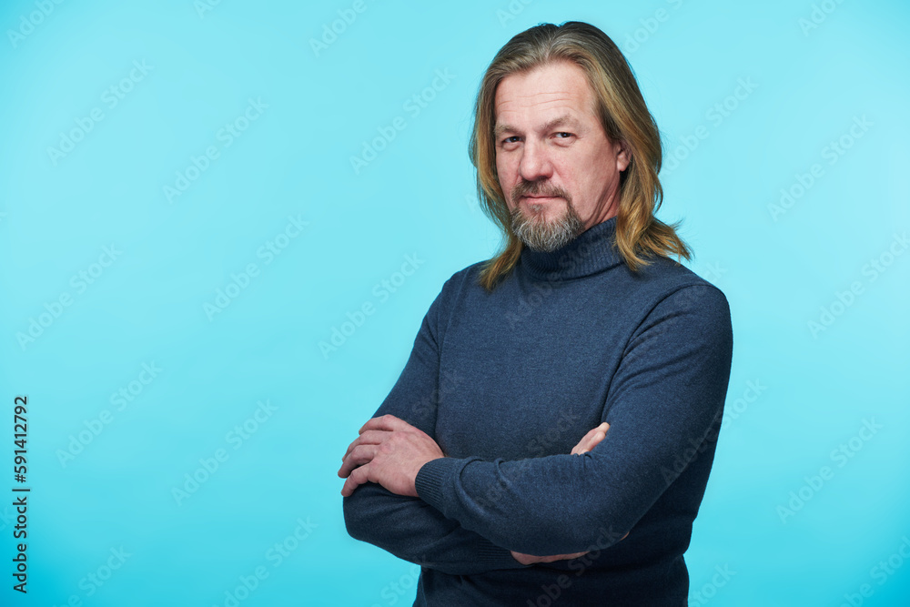 Fototapeta premium Portrait of mature bearded man in turtleneck looking at camera standing with his arms crossed on blue background