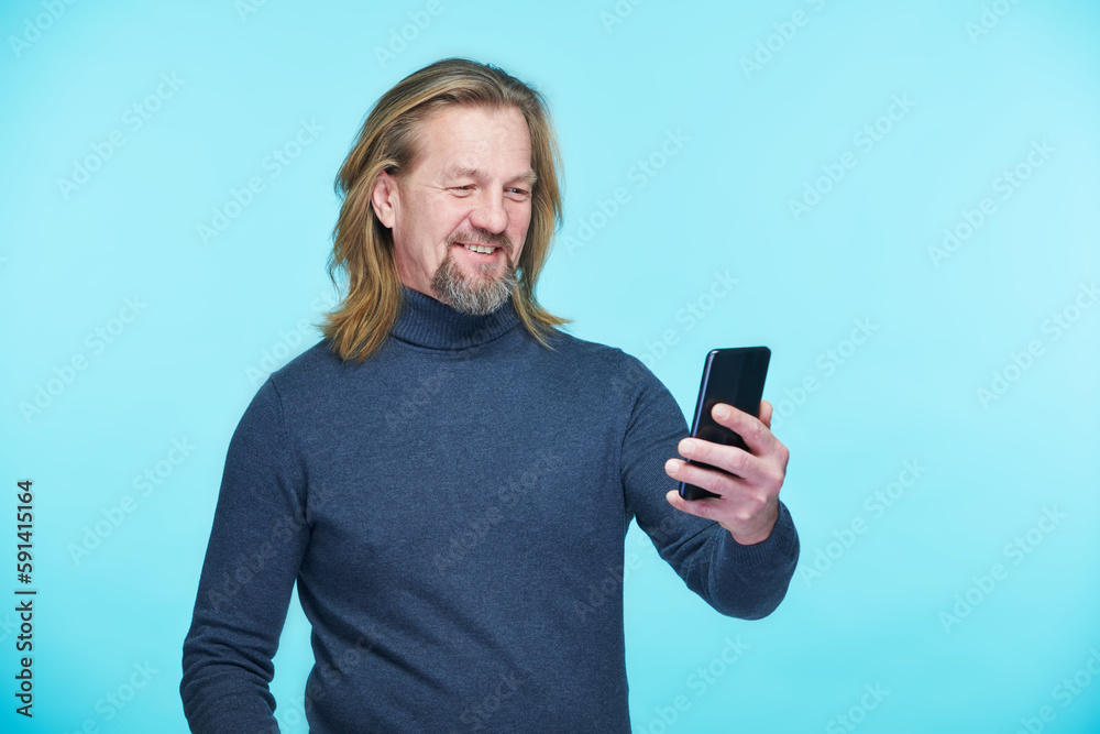 Mature man smiling during online communication on his smartphone standing against blue background Mature man smiling during online communication on his smartphone standing against blue background