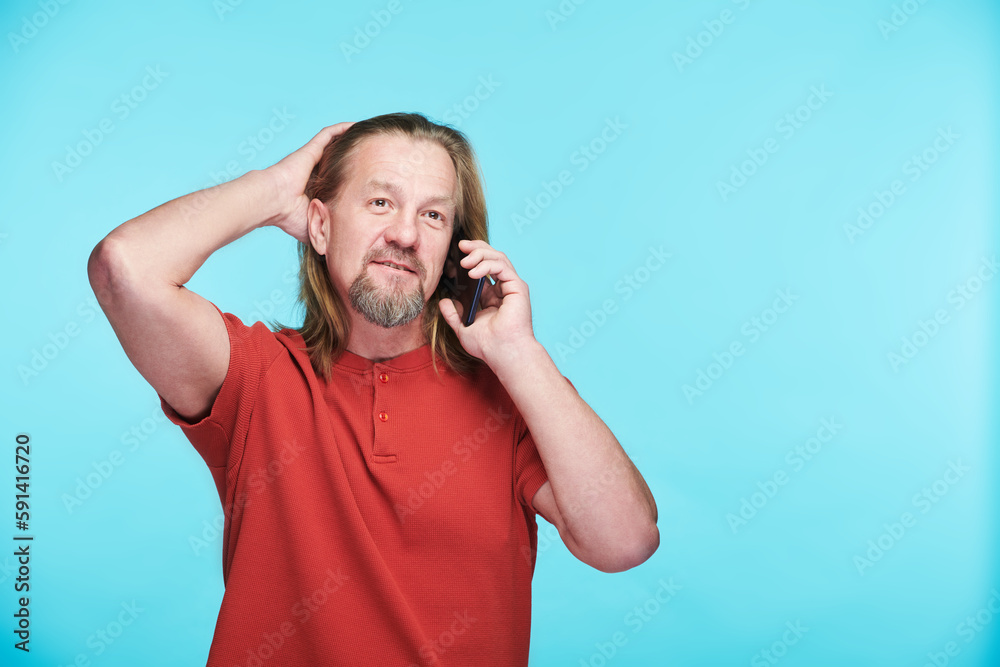 Mature man in red shirt having conversation on his mobile phone standing against blue background Mature man in red shirt having conversation on his mobile phone standing against blue background