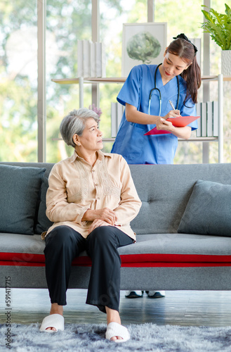 Asian professional successful female intern nurse in blue uniform with stethoscope standing talking asking symptoms from old senior elderly pensioner woman patient and writing note down in clipboard