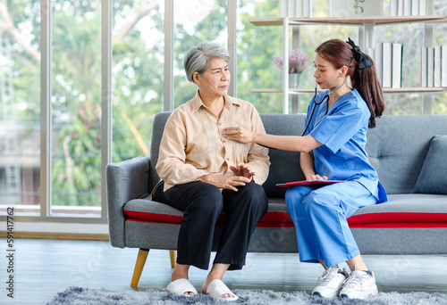 Asian professional successful female internship nurse in blue uniform sitting on sofa couch smiling using stethoscope listening to heart rate beating from old senior elderly pensioner woman patient