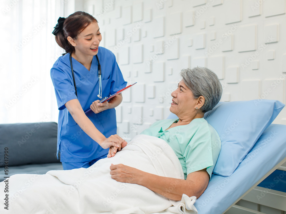 Asian professional successful female internship nurse in blue uniform with stethoscope visiting old senior elderly pensioner woman patient laying lying down on bed writing symptoms down on clipboard