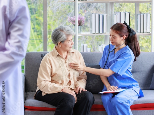 Asian professional successful female internship nurse in blue uniform sitting on sofa couch smiling using stethoscope listening to heart rate beating from old senior elderly pensioner woman patient